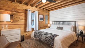 A wooden cabin bedroom with a bed, bedside tables, a chair, a mirror, and a window with curtains.