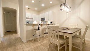 A modern kitchen with white cabinets, a stainless steel refrigerator, and a microwave. There is a dining area with a wooden table and white chairs.