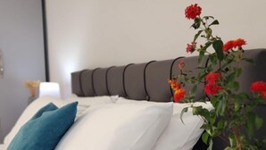En pænt redt seng med hvide sengetøj og en teal farvet pude, en grøn bog på et natbord, og en vase med røde blomster.