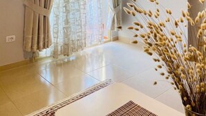 Et værelse med et stort vindue, beige gardiner, et billede i ramme på væggen og en vase med tørrede planter.