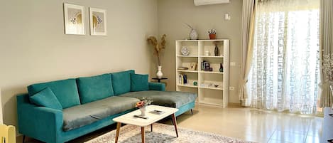 En stue med en turkis sofa, et sofabord, en bogreol og et stort vindue med gardiner.