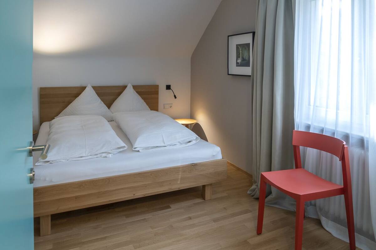 A bedroom with a bed, a chair, a bedside table, a wall-mounted lamp, and a window with curtains.