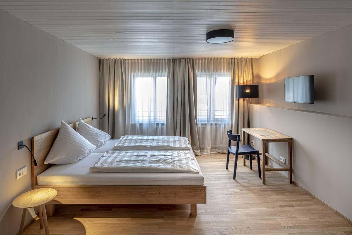 A bedroom with a wooden bed, a desk, a chair, a TV, and a window with curtains.