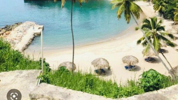 A beach with clear turquoise water, palm trees, and a white building in the background. - Wake Up At Paradise (Santa Barbara Samana)