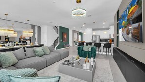 En moderne stue med en grå sofa, grønne lænestole og et sofabord. Der er en trappe, som fører til en etage ovenover, og et køkken er synligt i baggrunden.