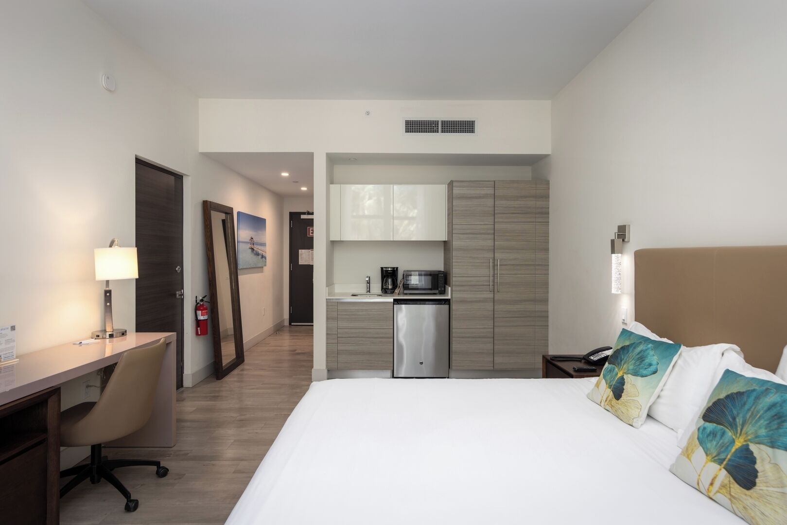 A hotel room with a bed, desk, chair, kitchenette, and a view of the hallway.