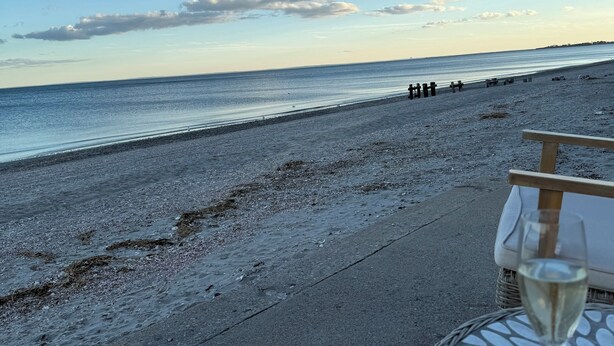Une vue sur la plage avec une table et un verre de vin, un banc en bois et une côte lointaine.
