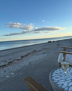 A beach view with a table and a glass of wine, a wooden bench, and a distant shoreline.