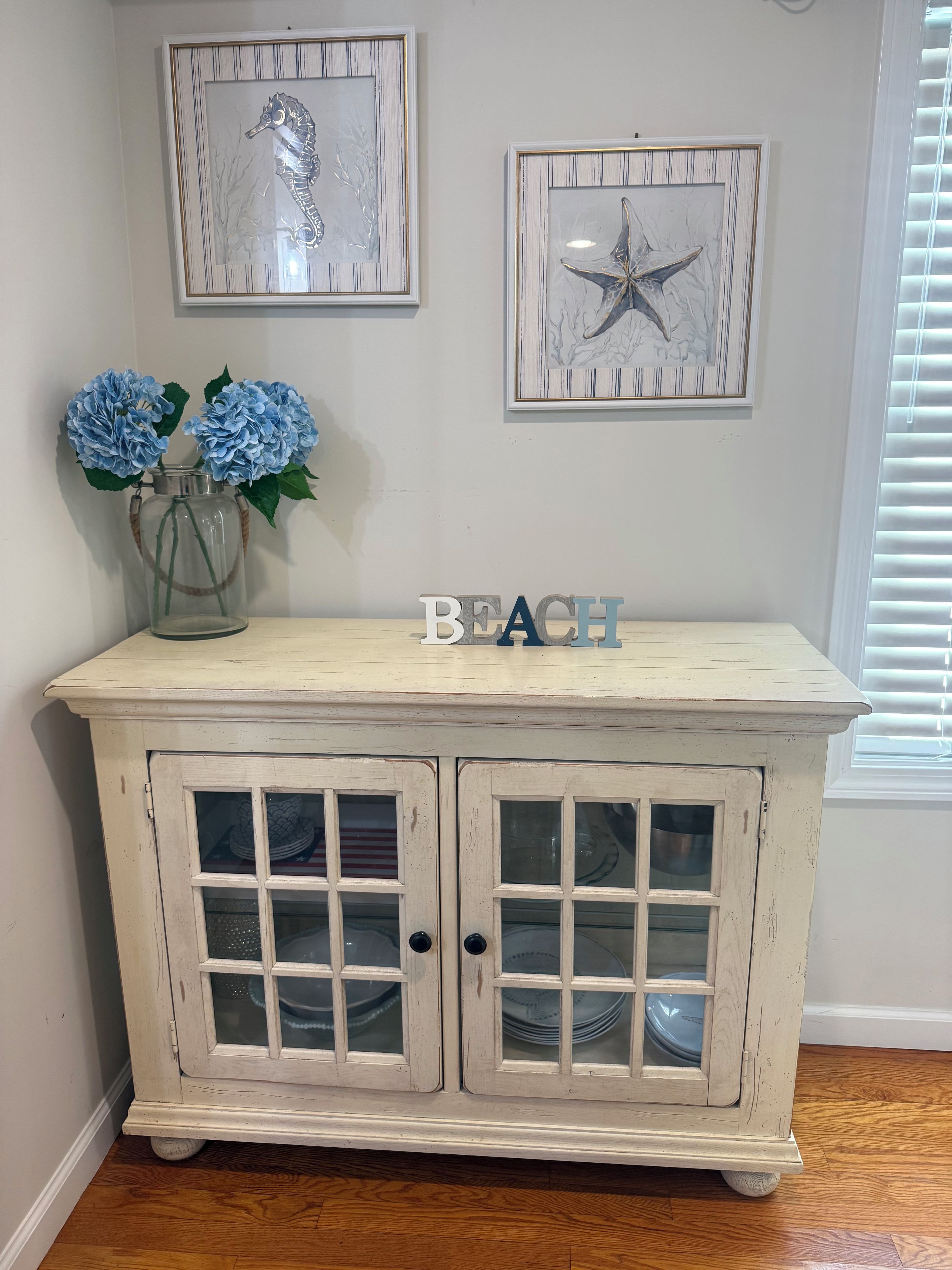 Un gabinete de madera blanca con puertas de cristal, un jarrón con flores azules y dos cuadros en la pared.
