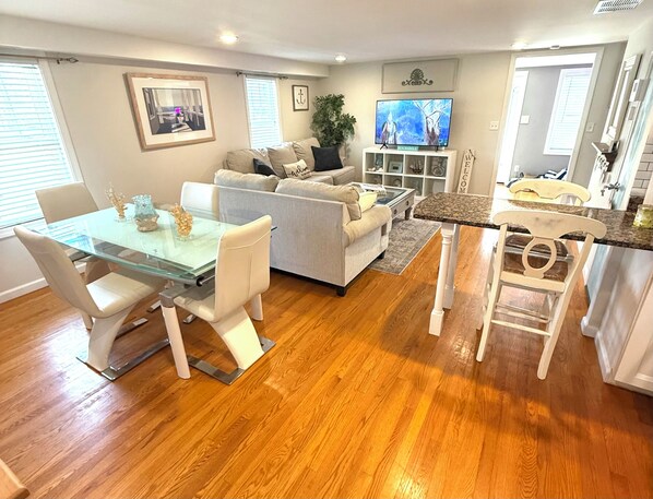 A modern living room with a glass dining table, white sofas, and a kitchen area with a granite countertop.