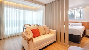 Una habitación de hotel moderna con sofá, cama, mesita de noche y ventana con vistas a edificios, además de una puerta corredera.