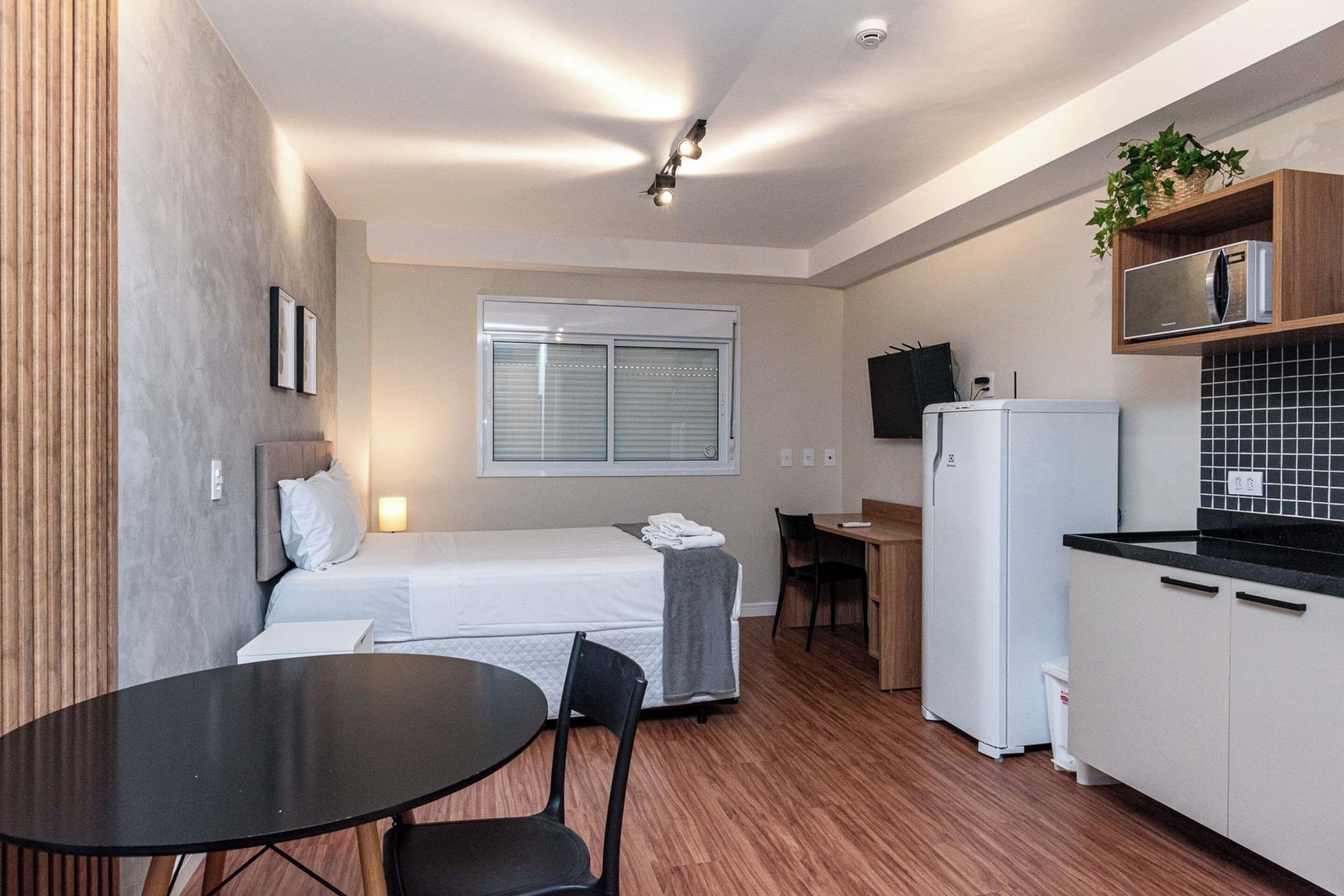 Une chambre d’hôtel de taille réduite, comprenant un lit, une petite table à manger, un coin cuisine et une fenêtre avec des stores.