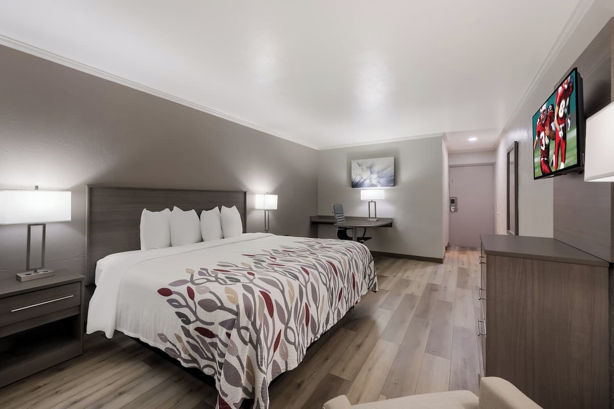 A hotel room with a large bed, bedside tables with lamps, a desk with a chair, a flat-screen TV mounted on the wall, and a painting above the desk.