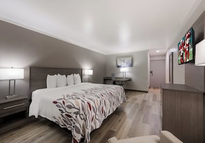 A hotel room with a large bed, bedside tables with lamps, a desk with a chair, a flat-screen TV mounted on the wall, and a painting above the desk. - Red Roof Inn Madisonville (Madisonville)