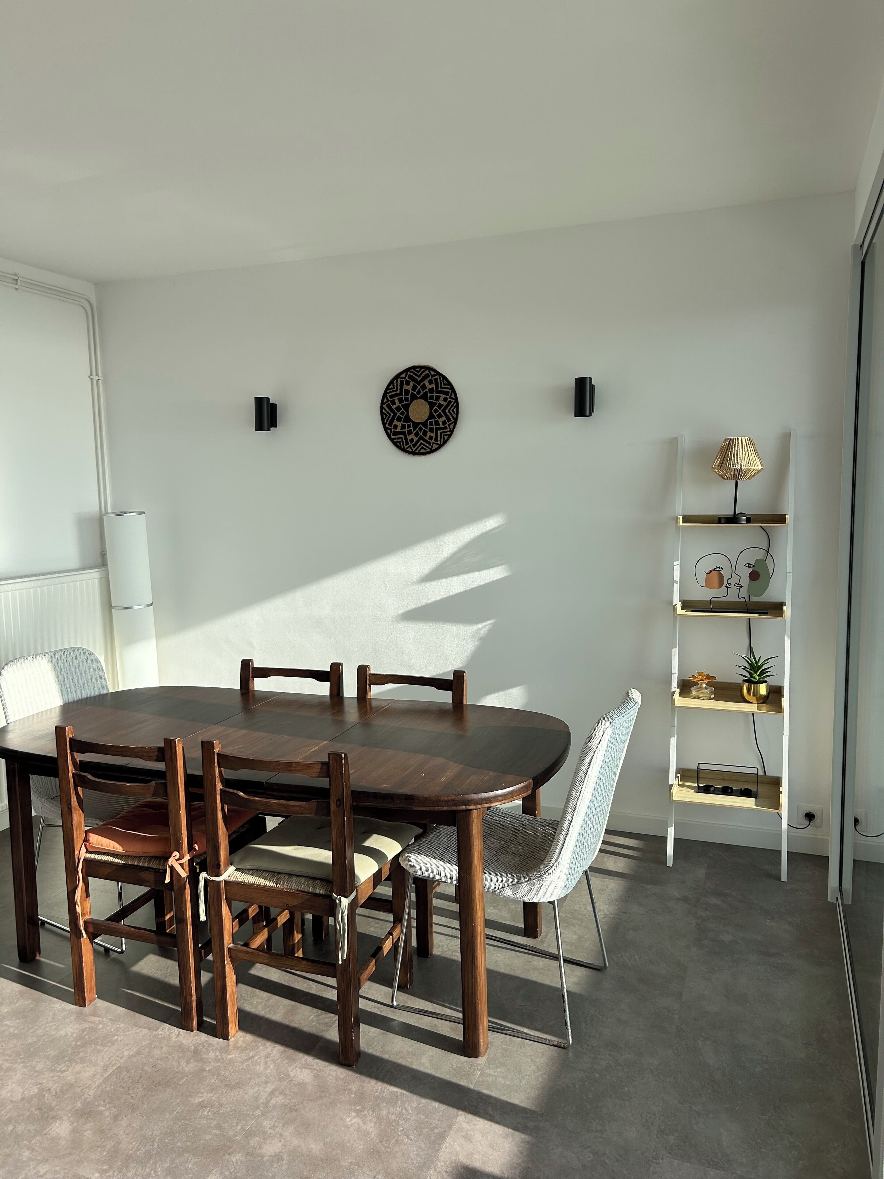 Un coin repas avec une table et des chaises en bois, un mur blanc orné d’une assiette décorative et une étagère blanche agrémentée d’objets décoratifs.
