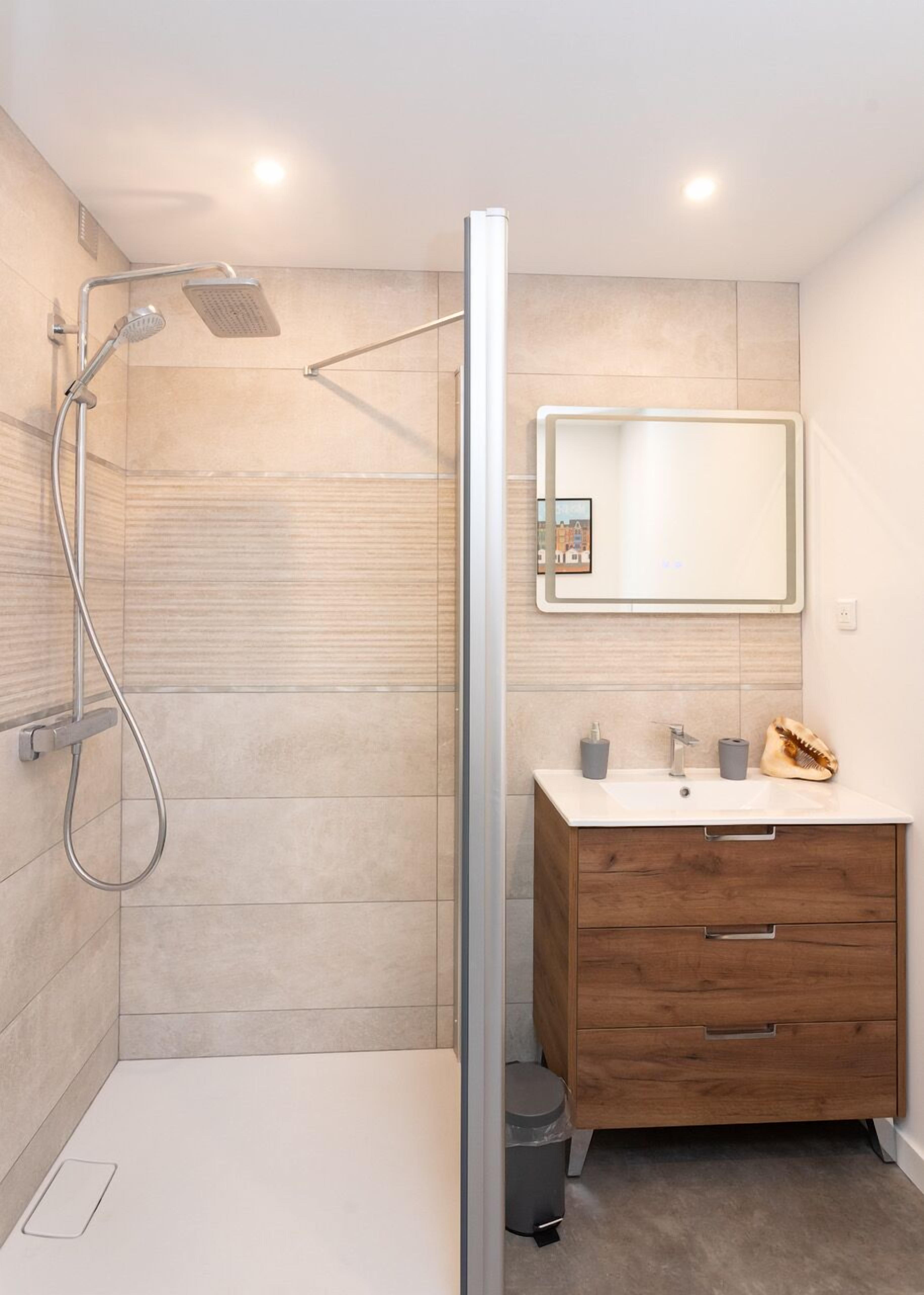 A modern bathroom with a glass shower enclosure, a wooden vanity with a mirror, and a small trash bin.