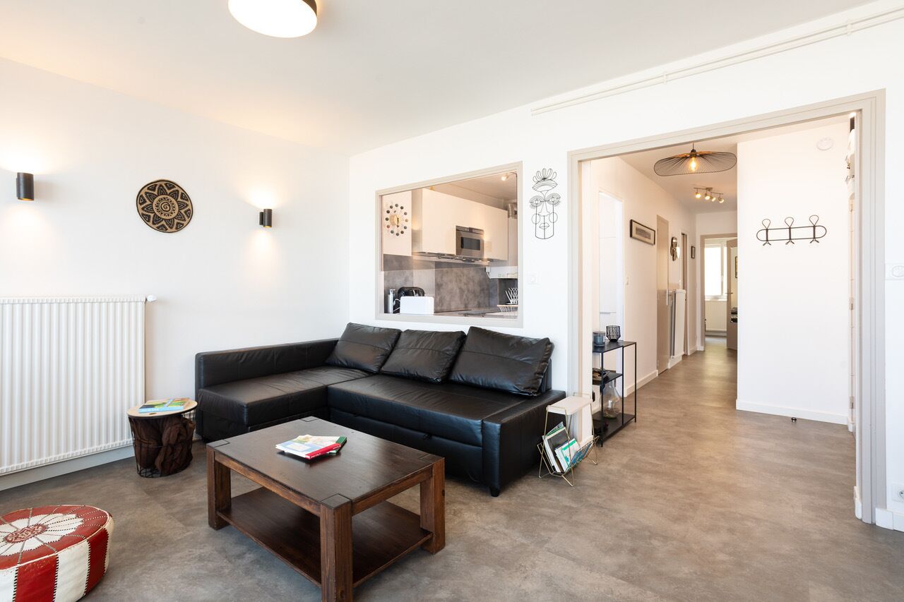 Un salon moderne avec un canapé d’angle, une table basse et une petite table d’appoint. On peut apercevoir la cuisine à travers une porte ouverte.