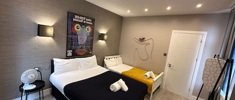 A modern hotel room with a large bed, a smaller bed, and a desk. There is a poster on the wall and a door with a towel rack.