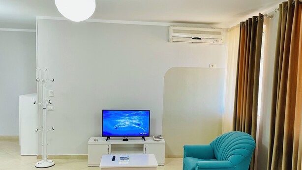 A modern living room with a flat-screen TV, a white cabinet, a teal armchair, and a white sofa with orange cushions. - EasyTrip Family - 2 bedrooms - City Center_417 (Divjakë)