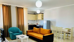 A living room with a sofa, a teal armchair, a coffee table, a dining table with chairs, and a kitchen in the background. - EasyTrip Family - 2 bedrooms - City Center_417 (Divjakë)