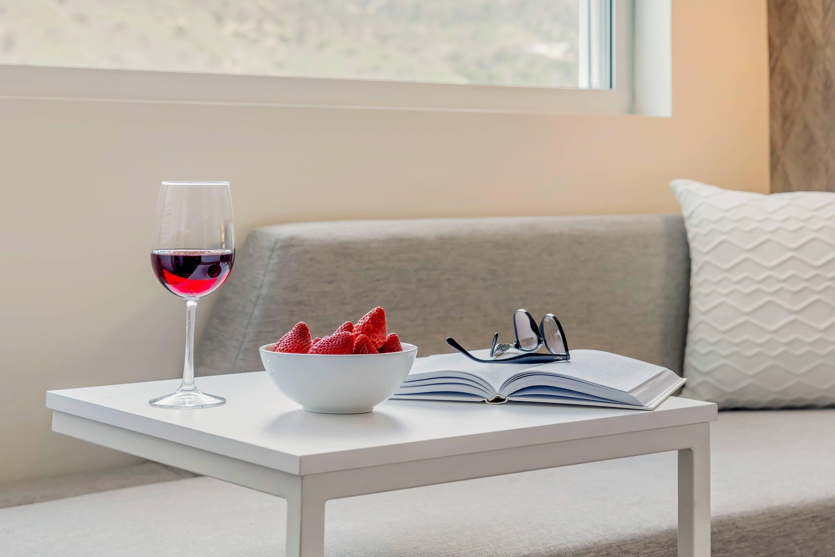 A white table with a wine glass, a bowl of strawberries, and an open book.