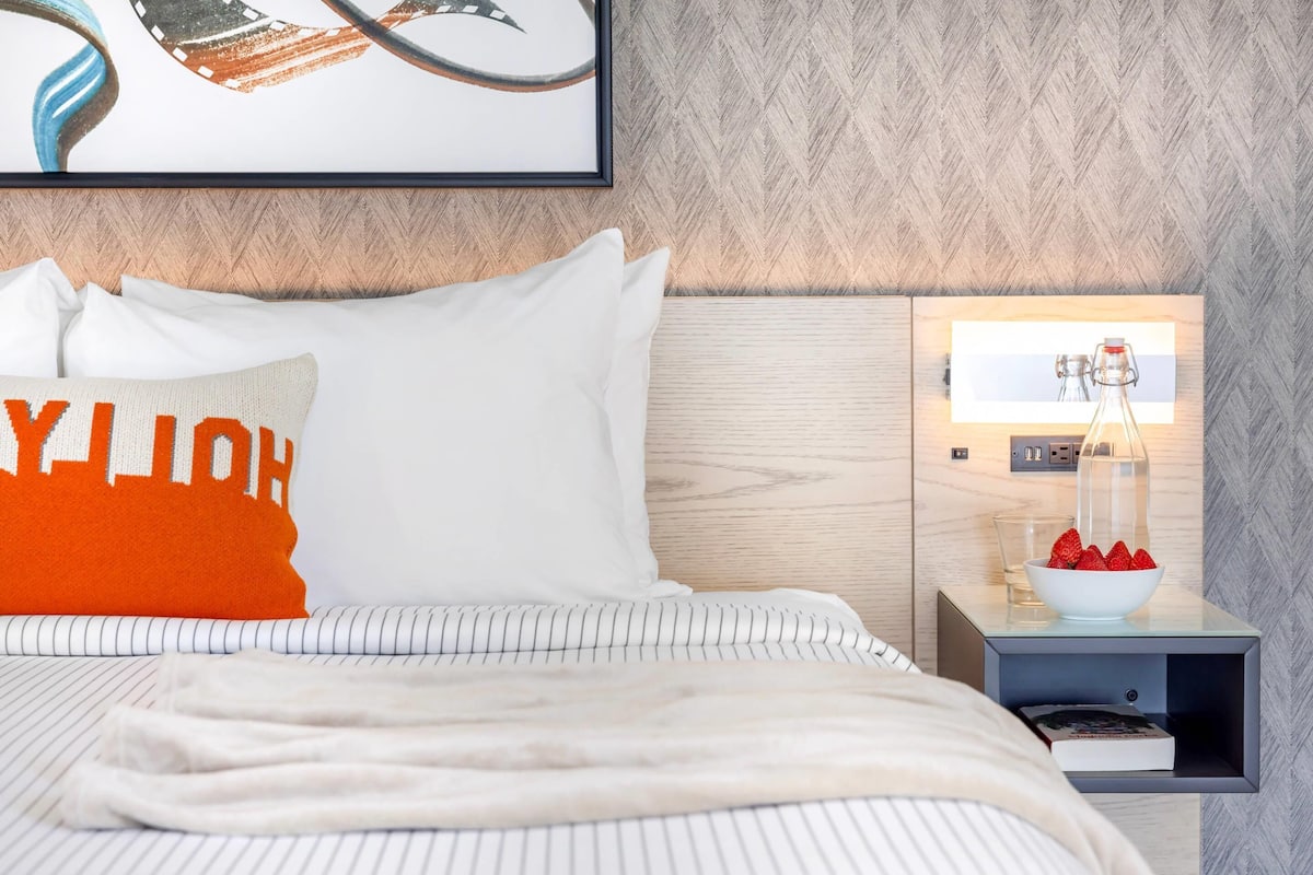 A neatly made bed with striped bedding, a white pillow with orange text, and a nightstand with a glass jar and strawberries.