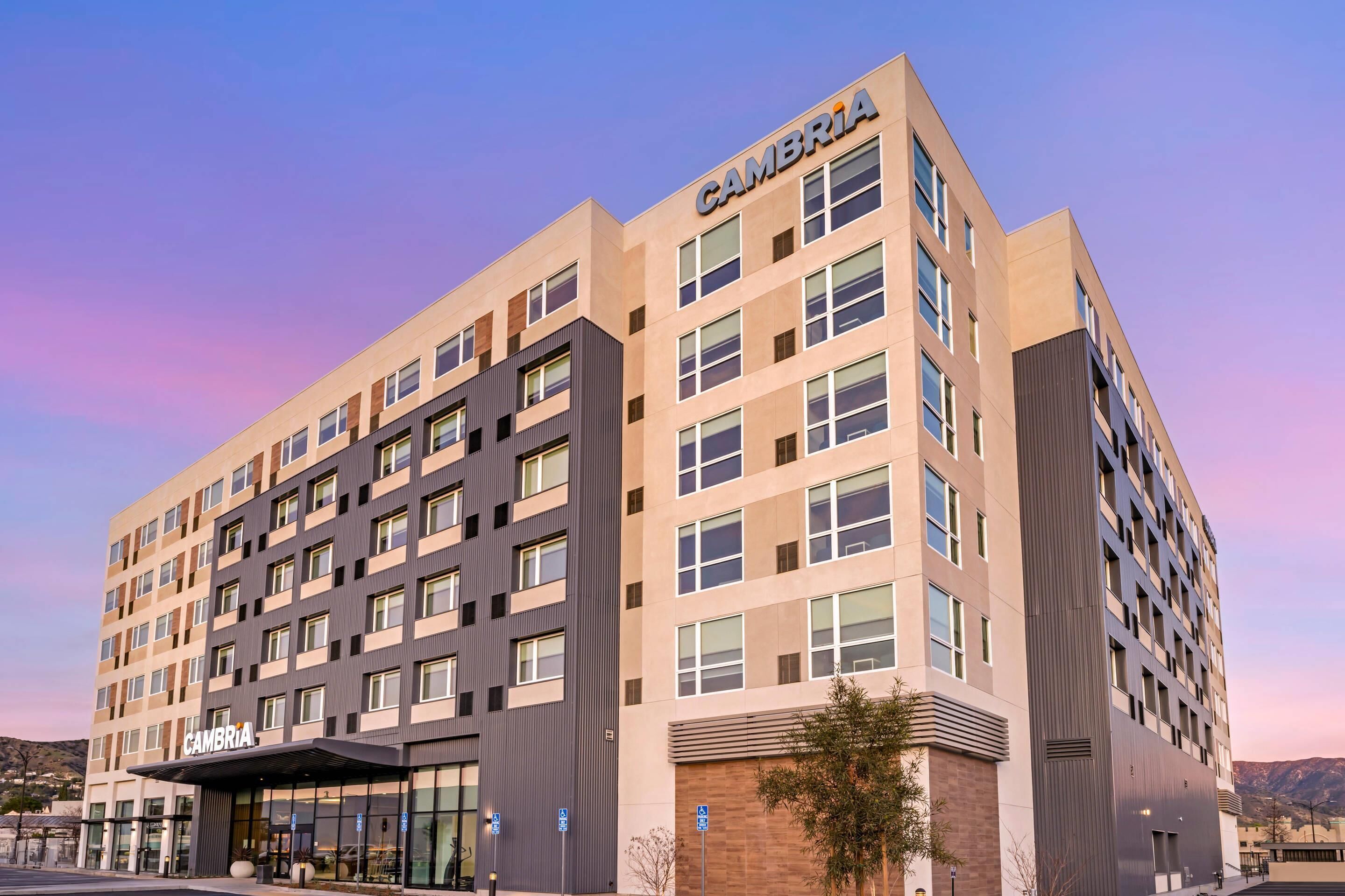 A multi-story hotel with the name 'CAMBRIA' on the facade, featuring a modern architectural design with a mix of window patterns and a flat roof.