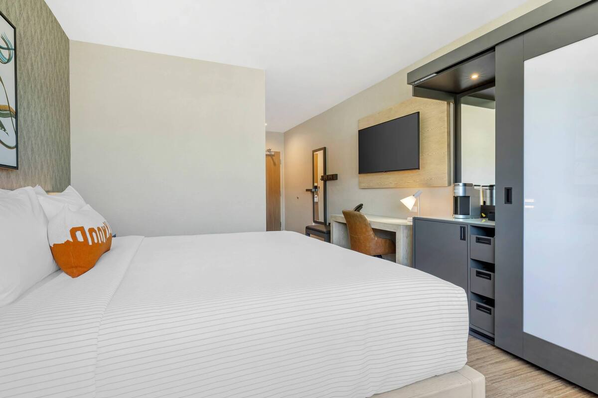 A modern hotel room with a large bed, a desk with a chair, a flat-screen TV mounted on the wall, and a minibar.