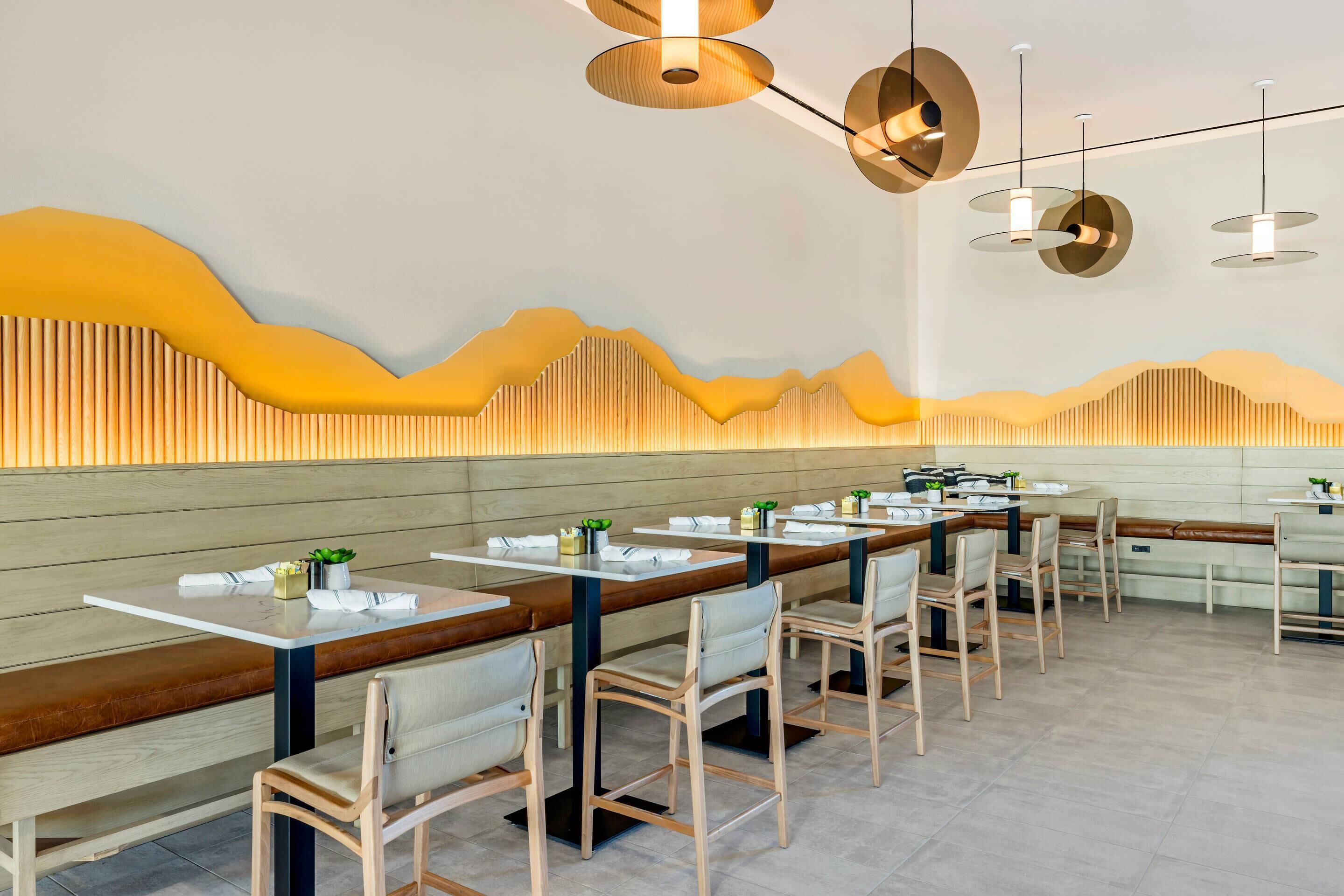 Un interior de restaurante moderno con mesas comunitarias largas, bancos acolchados e iluminación contemporánea.