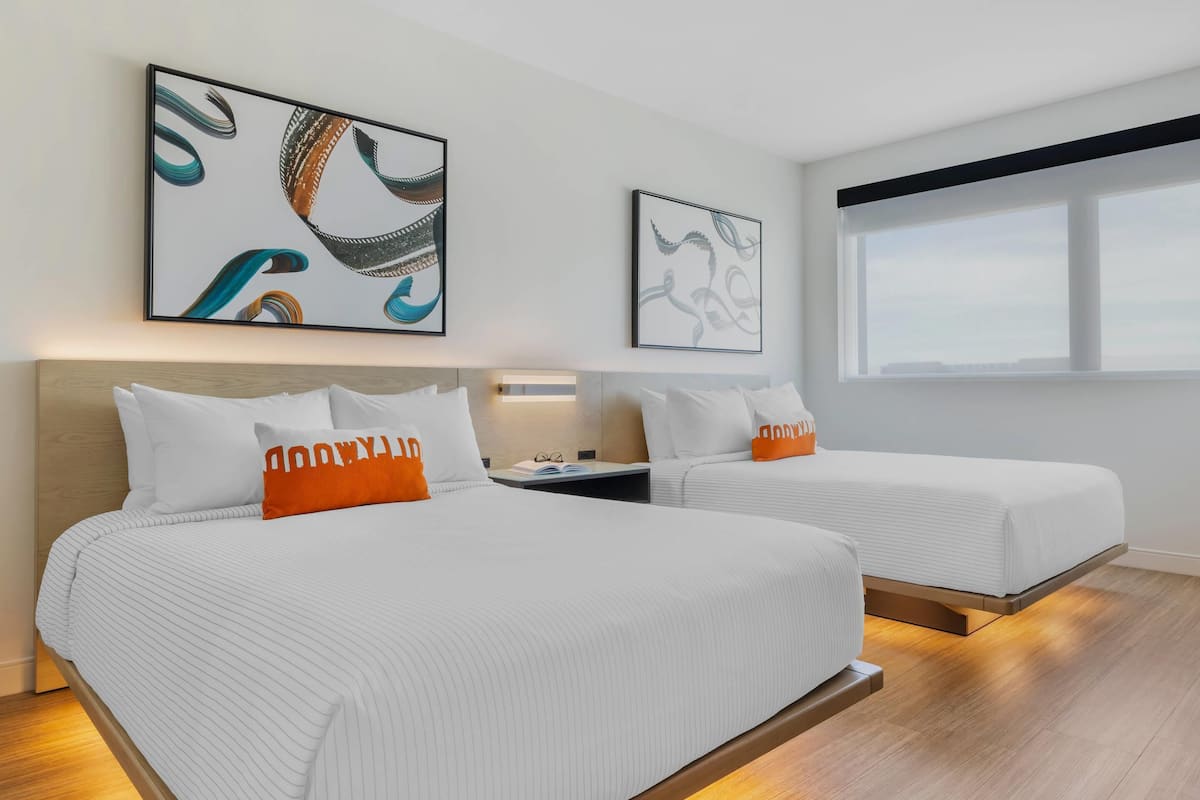 A hotel room with two beds, each with white bedding and orange pillows, a nightstand, and wall art.
