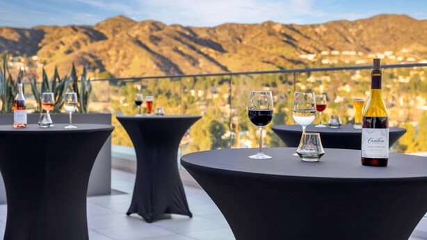 Una terraza con mesas redondas, cada una con una botella de vino y copas, con vistas a un paisaje montañoso.