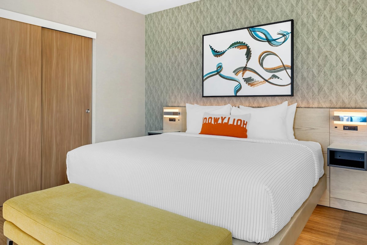 A hotel room with a large bed, a bench, a wardrobe, and a wall-mounted artwork.