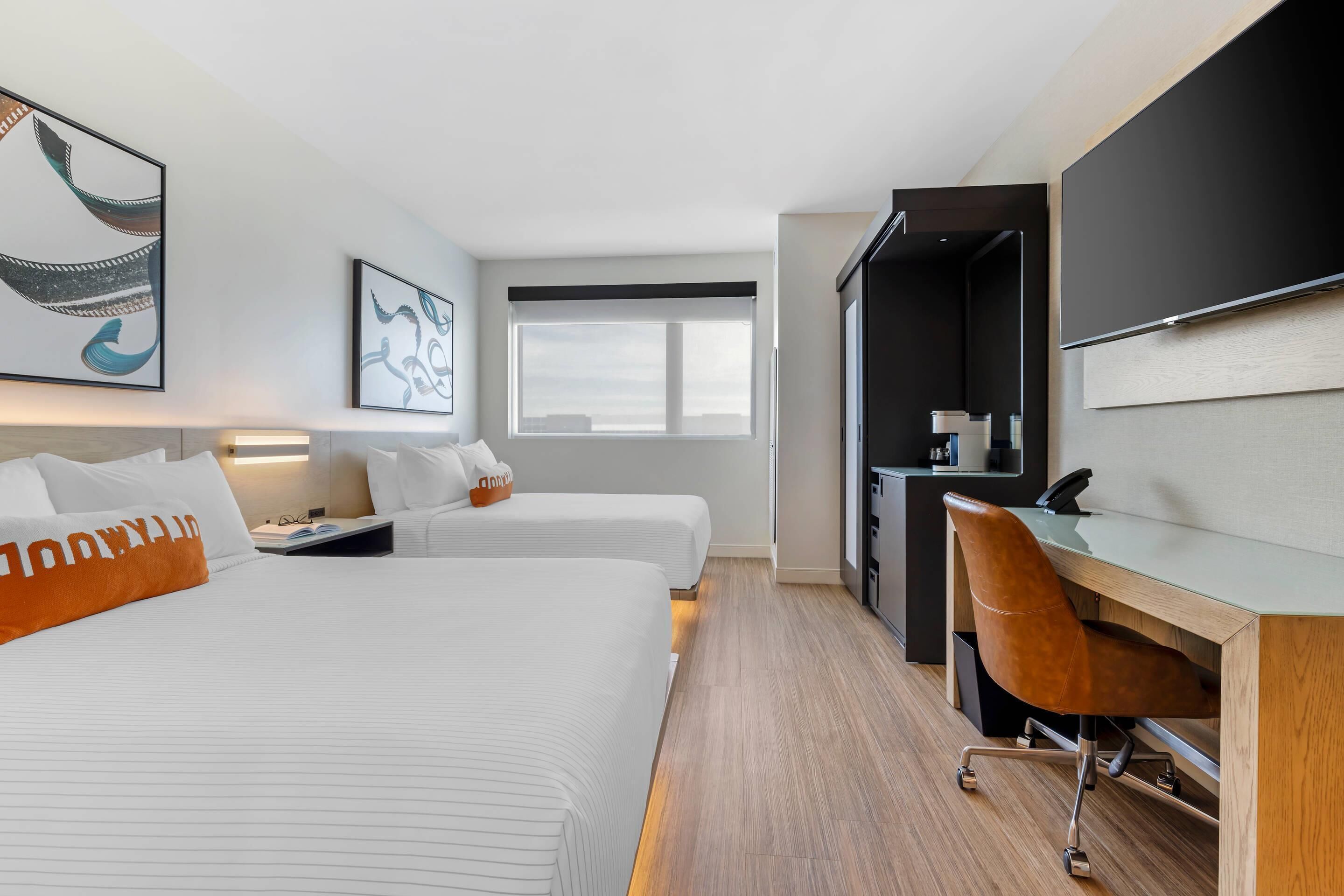 Quarto de hotel com duas camas, uma escrivaninha, uma cadeira, uma janela grande e obras de arte nas paredes.