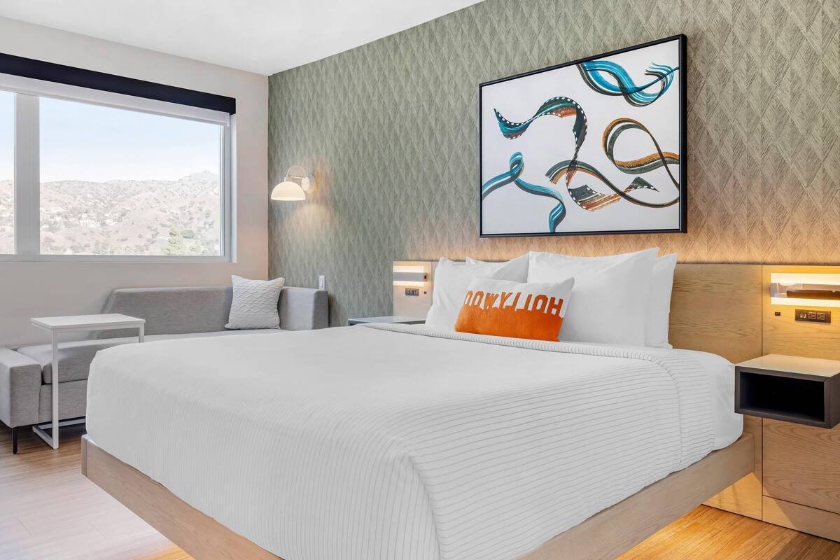 A modern hotel room with a large bed, a view of the mountains, and a wall-mounted abstract painting.