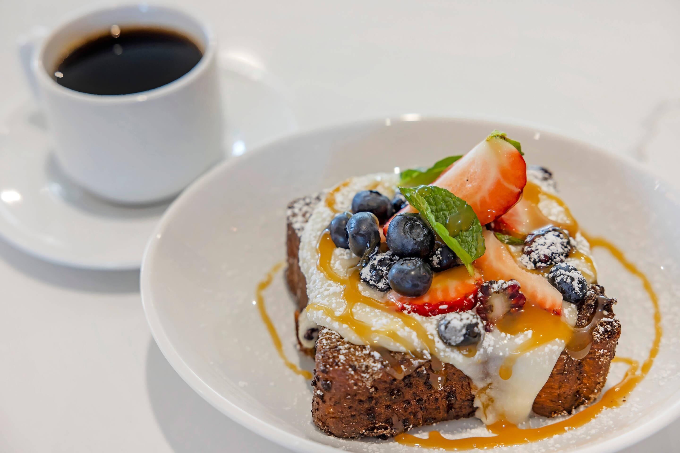 A dessert plate with a slice of cake topped with whipped cream, blueberries, strawberries, and a mint leaf, accompanied by a cup of coffee.