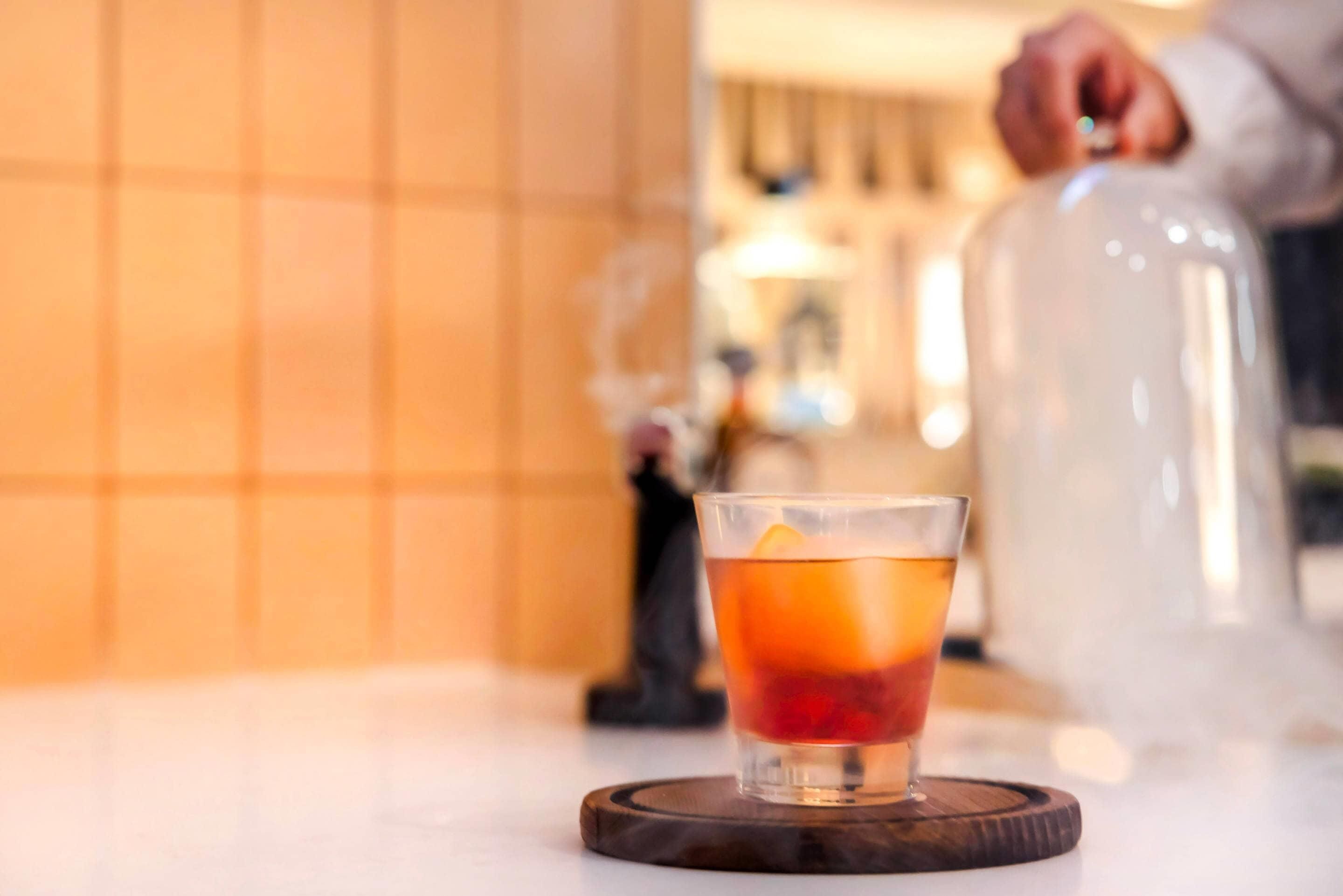 Ein Glas bernsteinfarbenes Getränk auf einem Holzuntersetzer, im unscharfen Hintergrund eine Person und ein großer Behälter.