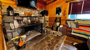 Ein Interieur einer Blockhütte mit Kamin aus Stein, einem darüber montierten Fernseher, einem Fenster mit Jalousien und einem braunen Ledersofa mit Kissen.