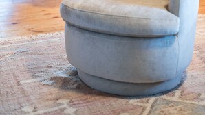 Een kamer met een grijze gestoffeerde fauteuil, houten wanden, een eettafel met stoelen en een raam met jaloezieën.