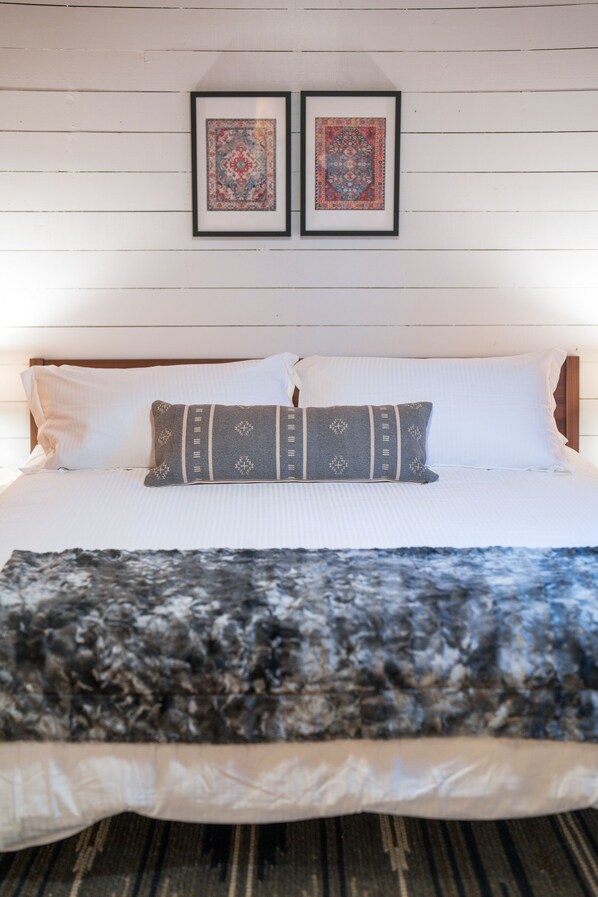 Un lit bien fait, avec du linge de lit blanc et un coussin à motifs gris, un tapis encadré sur le sol et deux autres tapis encadrés au mur.
