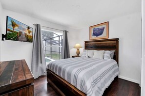 Une chambre à coucher avec un lit en bois, un téléviseur fixé au mur, une fenêtre avec des rideaux et un tableau encadré au-dessus du lit.