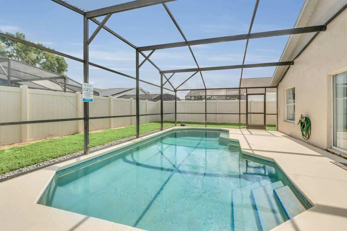 A rectangular swimming pool with a glass enclosure, surrounded by a white fence and a grassy area.