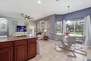A modern kitchen with a central island, dining area with chairs, and a TV on the wall.