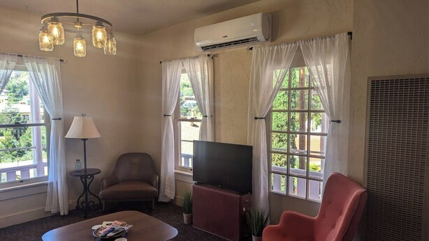 A living room with a sofa, a red armchair, a TV, and a coffee table. There are two windows with curtains, an air conditioning unit, and a chandelier.
