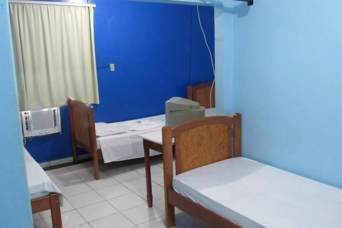 A small, single-room accommodation with a blue wall, a wooden bed, a wooden chair, a small table with a white tablecloth, and a window with a curtain.