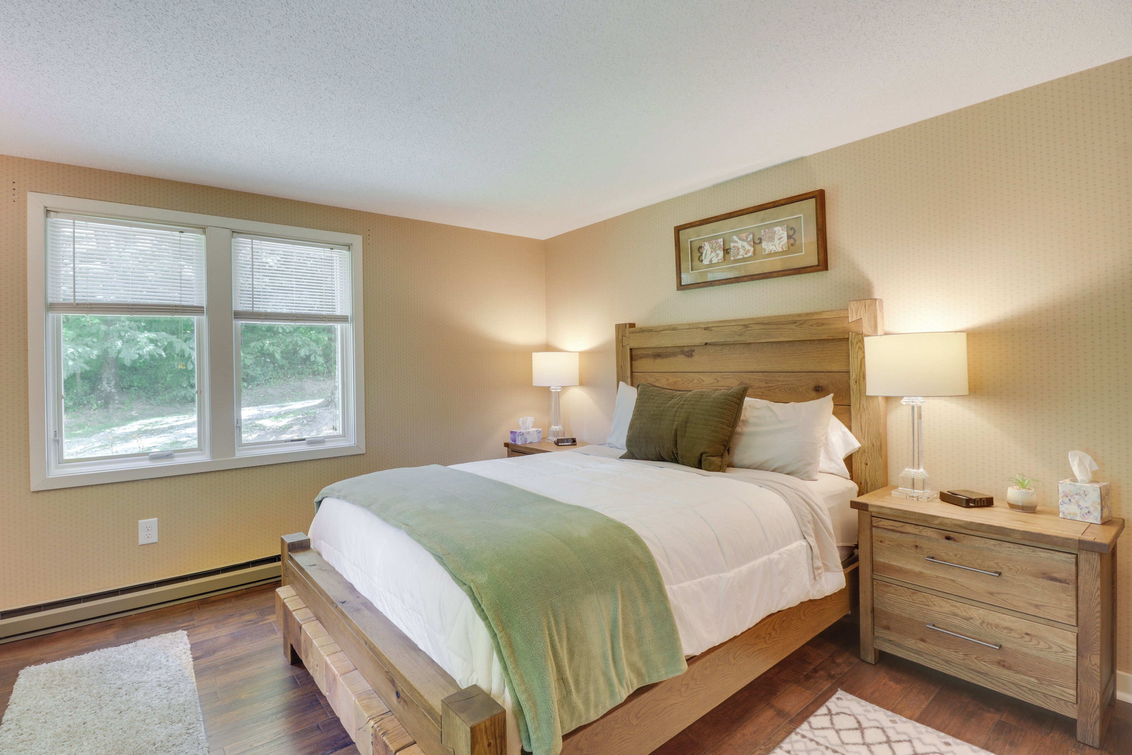 A bedroom with a wooden bed, two bedside tables with lamps, a nightstand, a framed picture on the wall, and a window with blinds.