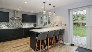 Une cuisine moderne dotée d’un grand îlot central, de tabourets de bar, de luminaires suspendus et offrant une vue sur le jardin par la porte.
