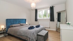 Une chambre avec une tête de lit bleue, un lit avec une literie grise, une table de chevet avec une lampe, une fenêtre avec des rideaux, un radiateur, un miroir et une commode blanche.