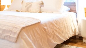 A bedroom with a wooden floor, a bed with white bedding, a patterned rug, and a window with curtains.