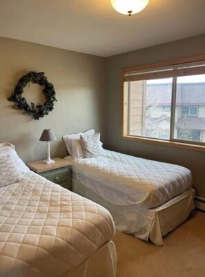 Two single beds with quilted mattresses and white bedding, a nightstand with a lamp, and a large window with a residential view.