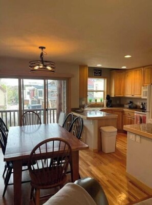 A modern kitchen with wooden cabinets, a dining area with a table and chairs, and a view of the outside through large windows. - Walkable Winter Park Oasis! (Winter Park)